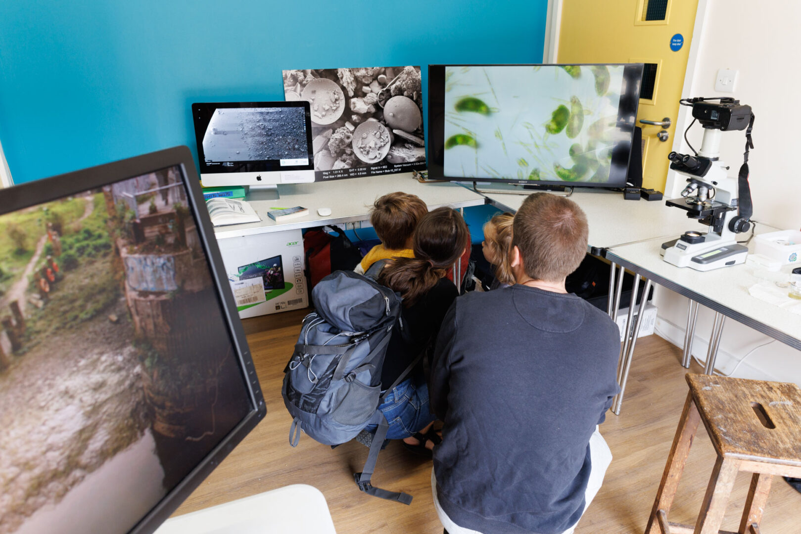 children and adults cluster around microscope and flatscreen showing microalgae samples. Around them are other screens and posters